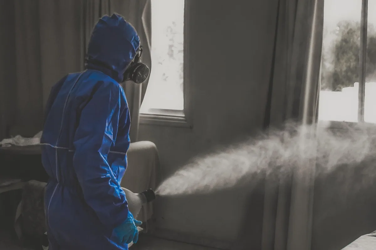 Aplicador con mono, guantes y máscara de protección nebulizando un producto en una habitación