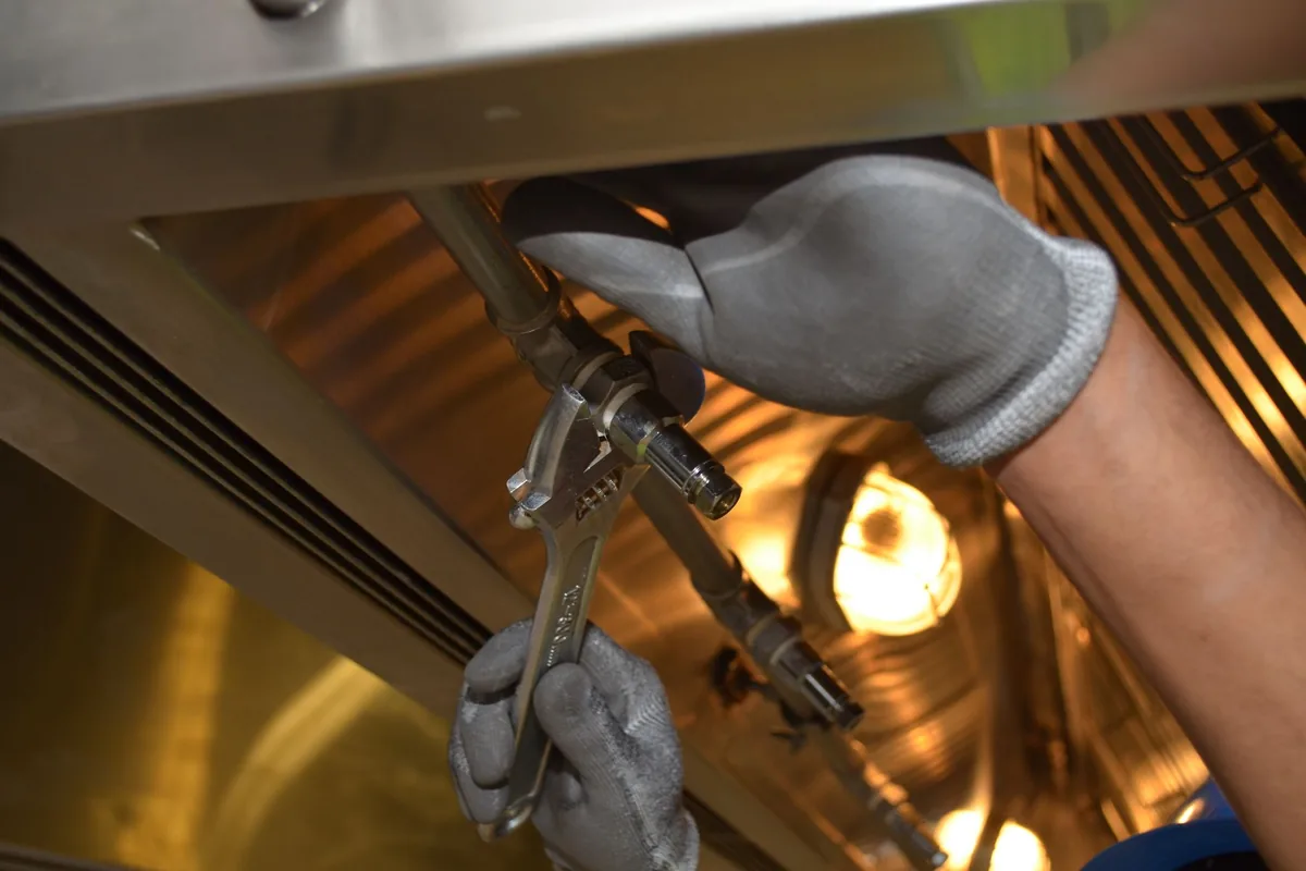 Manos con guantes ajustando con una llave inglesa la tuerca de un tubo bajo una campana extractora de acero inoxidable