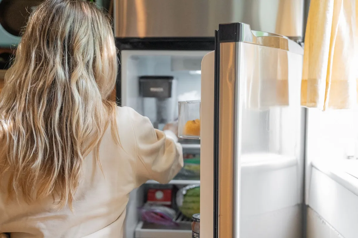 Frigorífico de acero inoxidable abierto, con las baldas y la comida a la vista, y una mujer de espaldas metiendo la mano